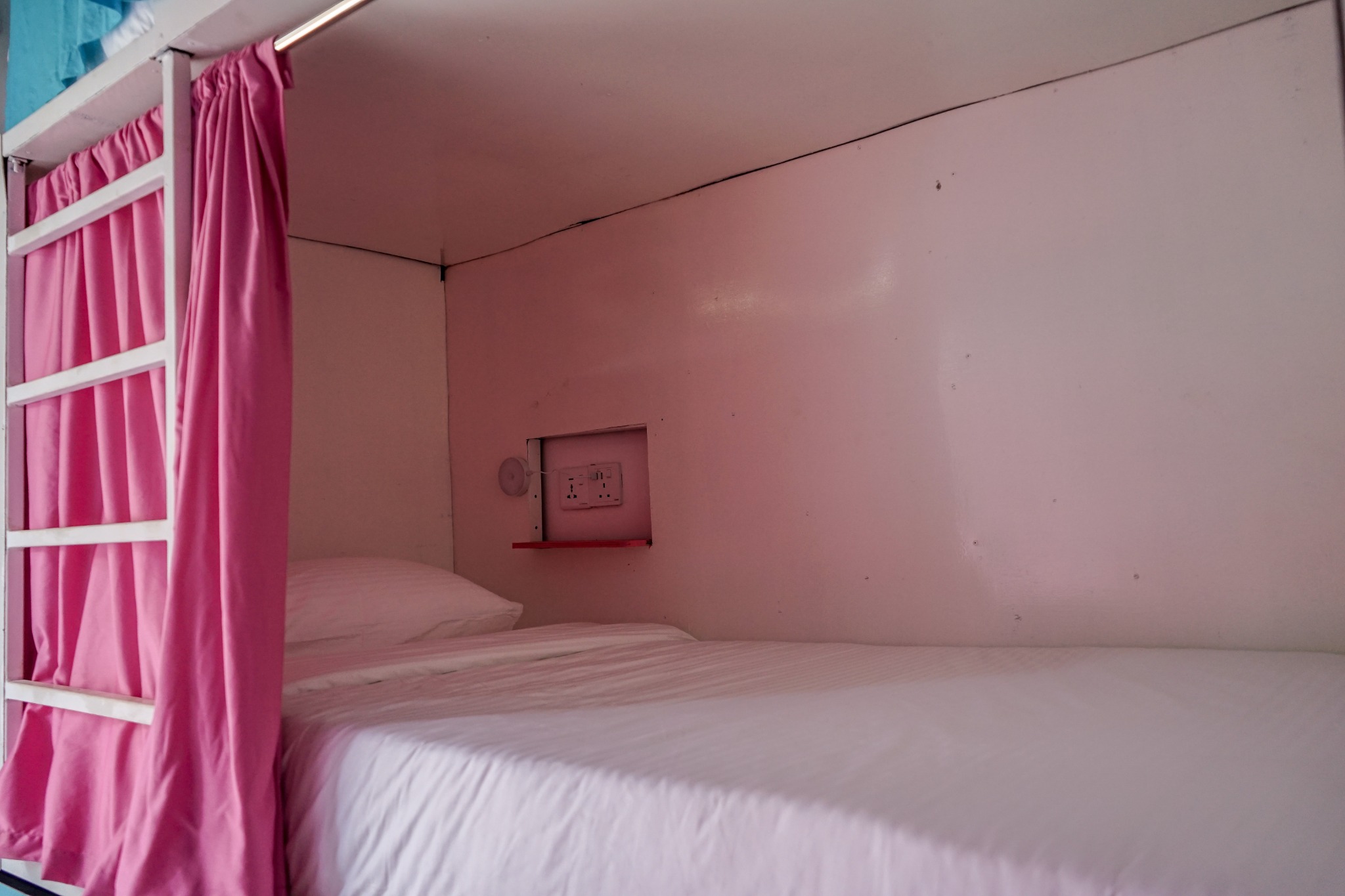 Dorm bed with white bedding, reading light and socket behind a pink curtain