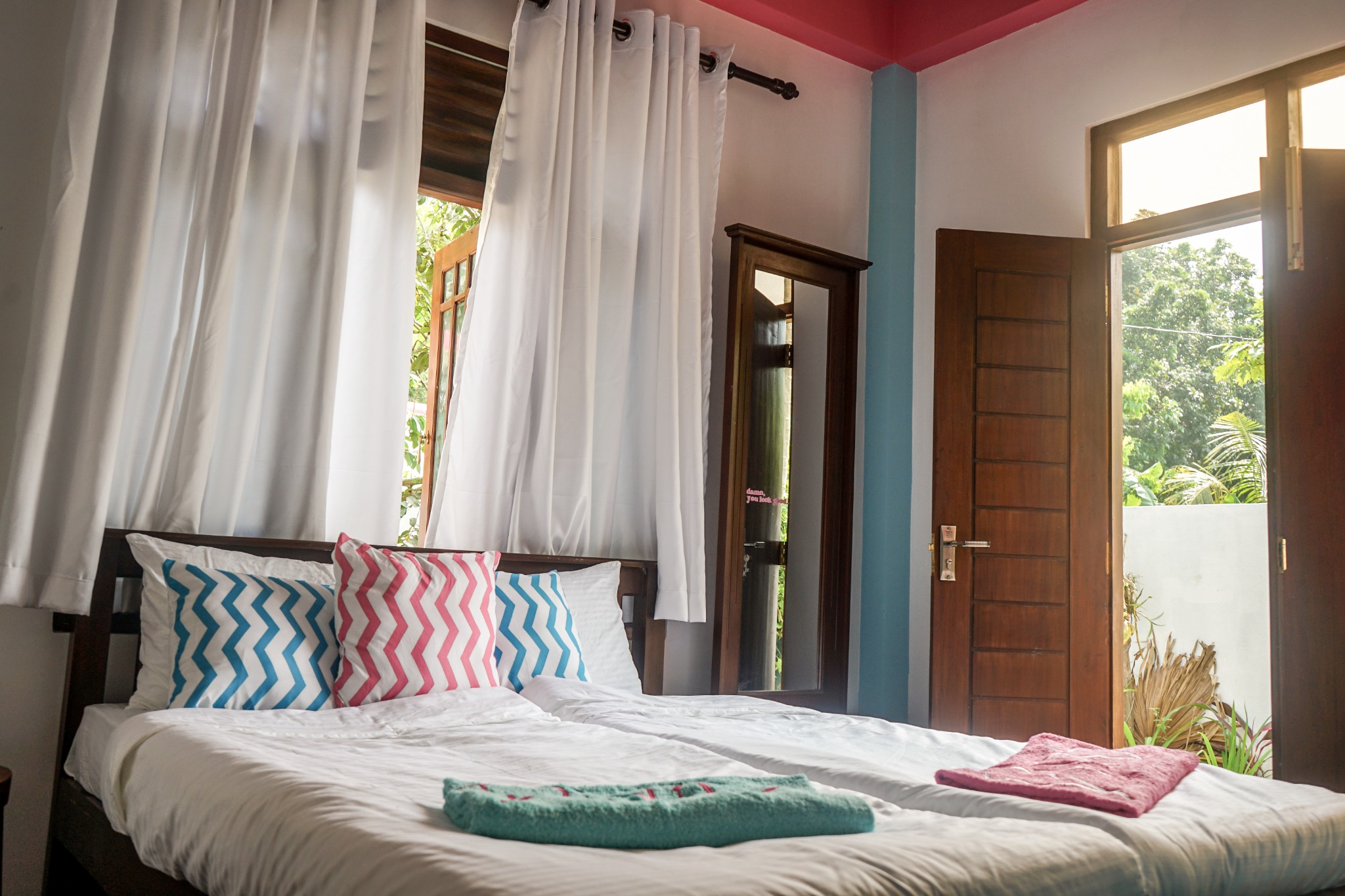 Private bedroom with two made beds, white curtains, wooden furniture and a door opening to greenery